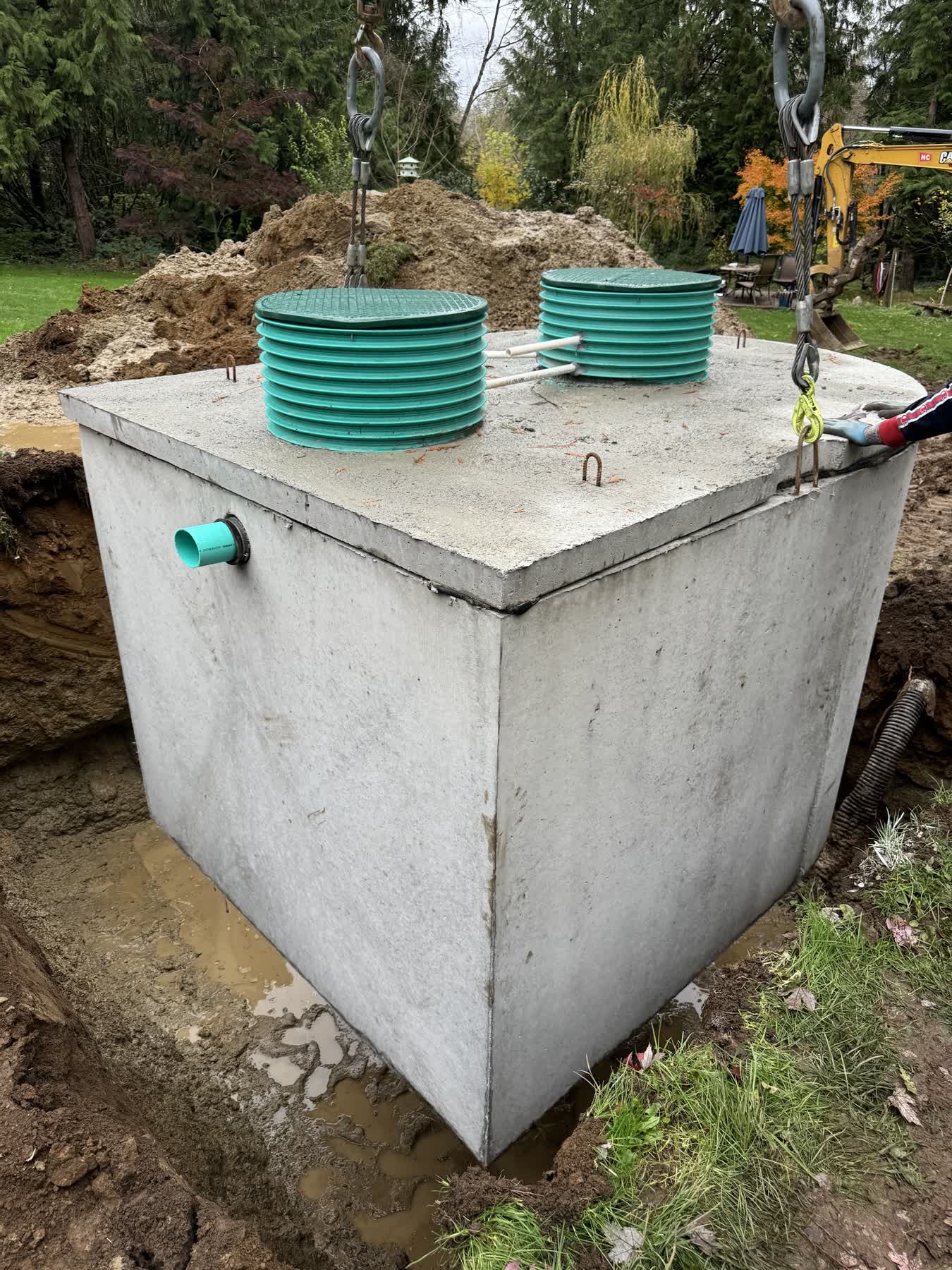 A precast concrete septic tank with two teal risers being lowered into place on a rural property.