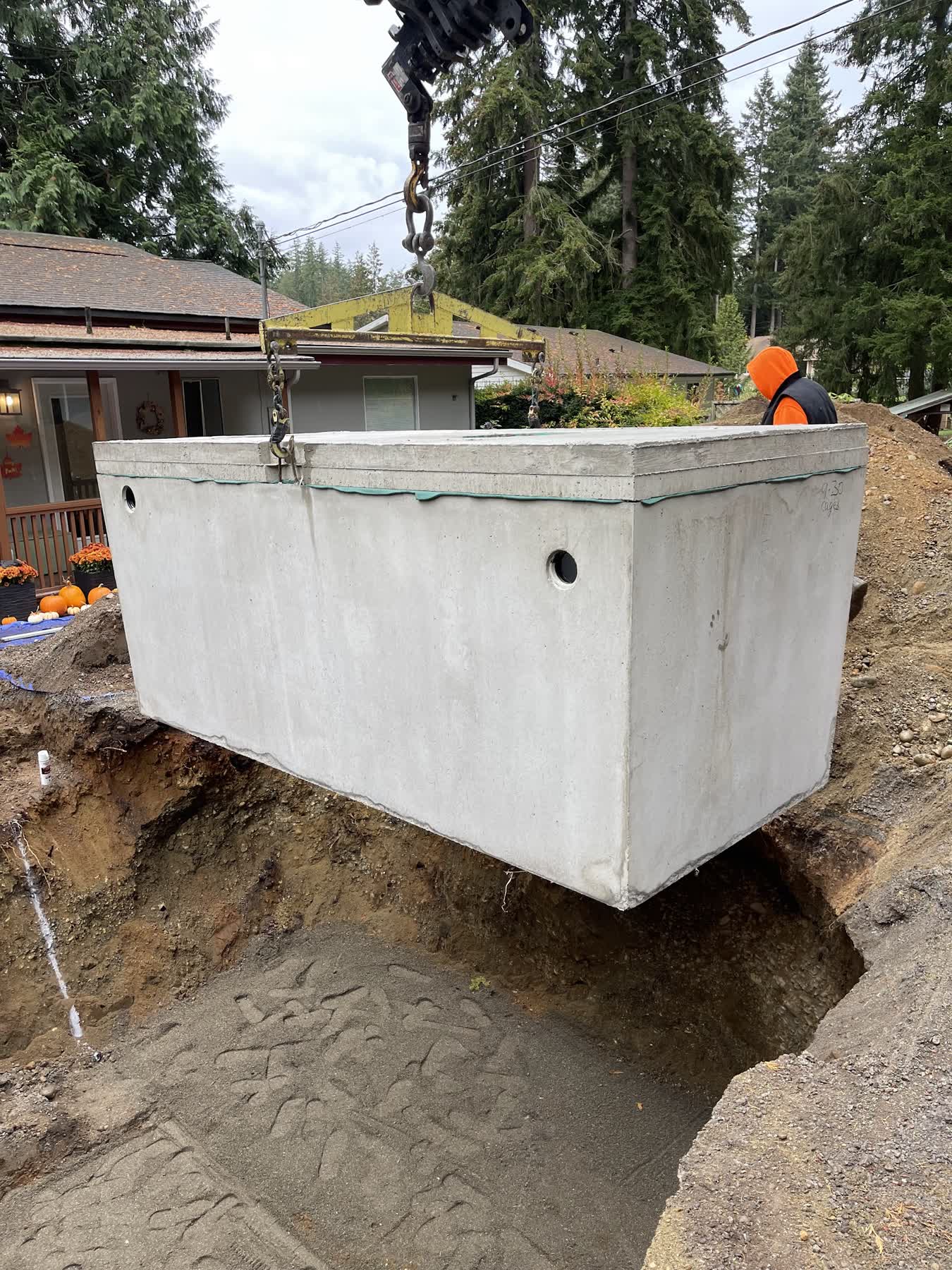 A crane lowering a large concrete septic tank into a prepared pit beside a home.