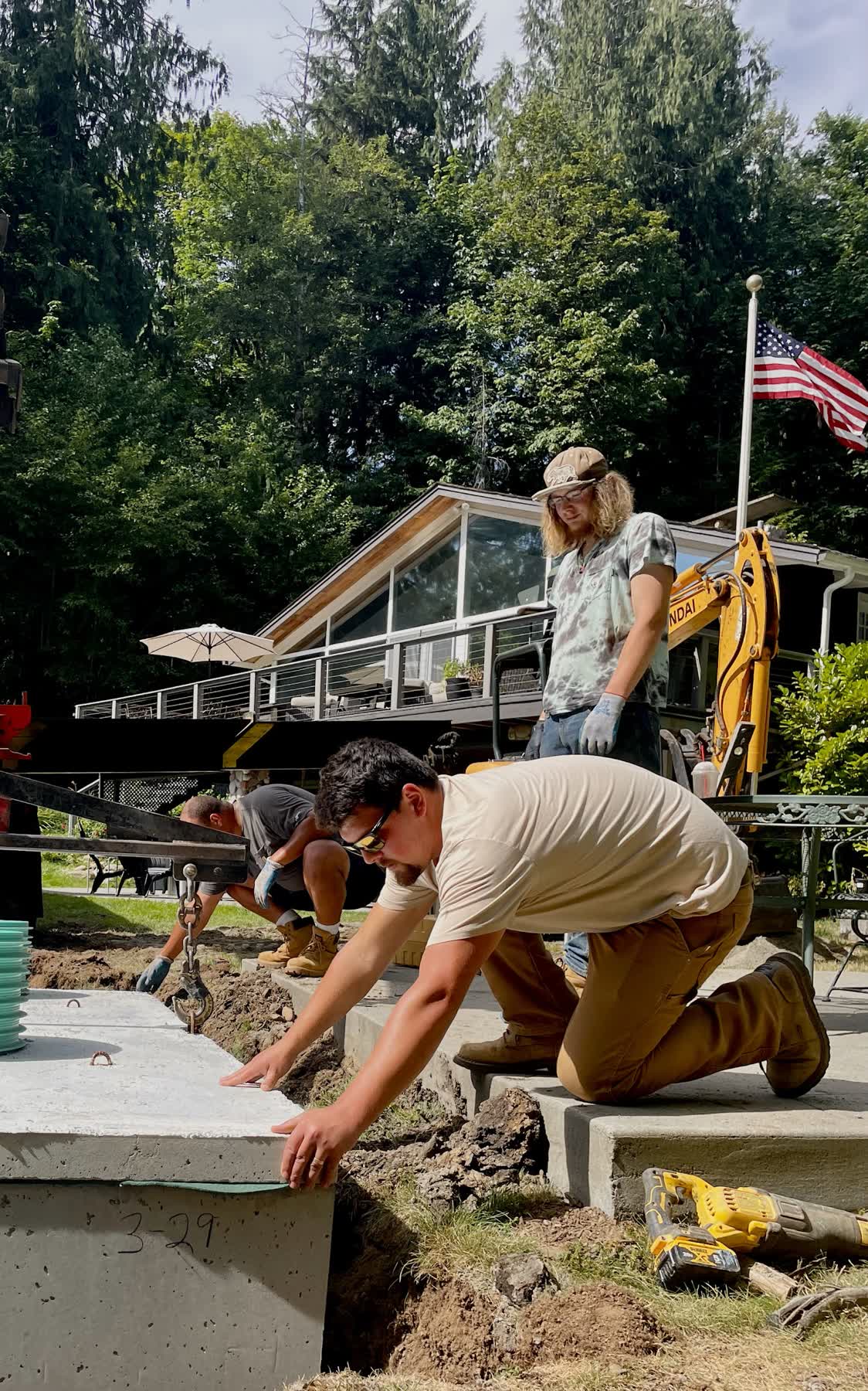 Two crew members setting a concrete septic tank lid at a lakeside home, with a flag flying behind them.