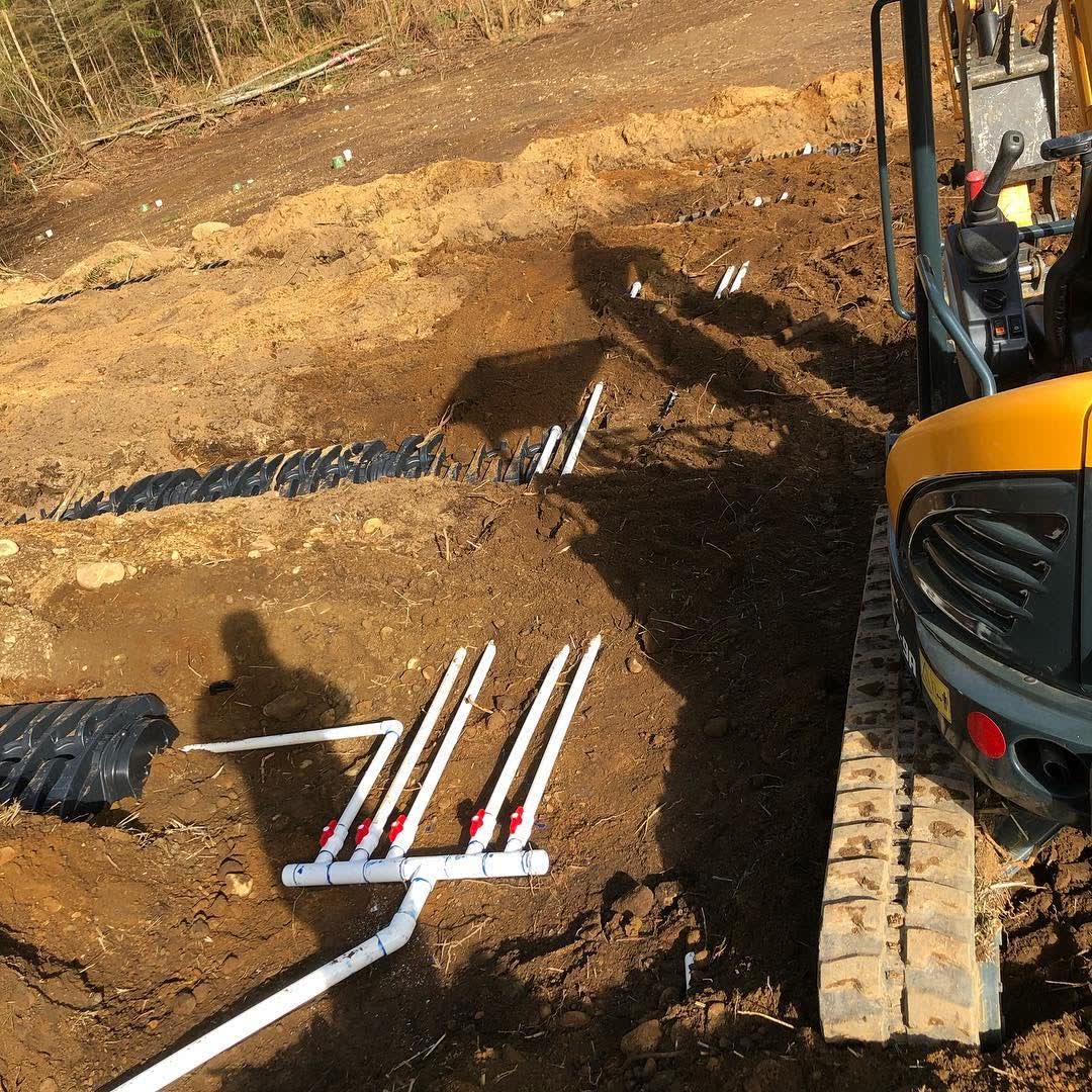Drain field chambers and white distribution pipes laid in a graded trench next to a mini excavator.