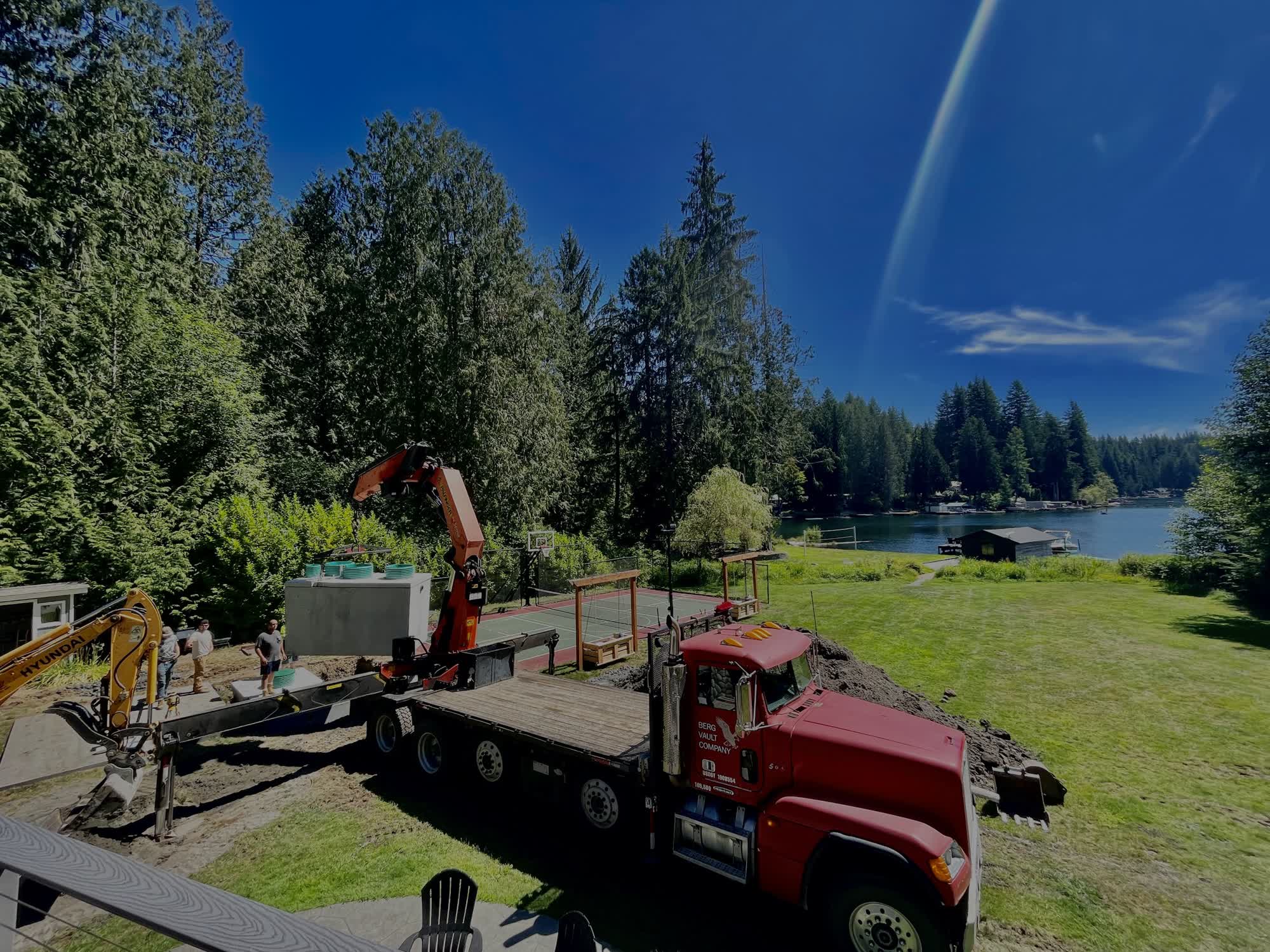 A crane truck setting a concrete septic tank beside a lakefront yard surrounded by evergreen trees.