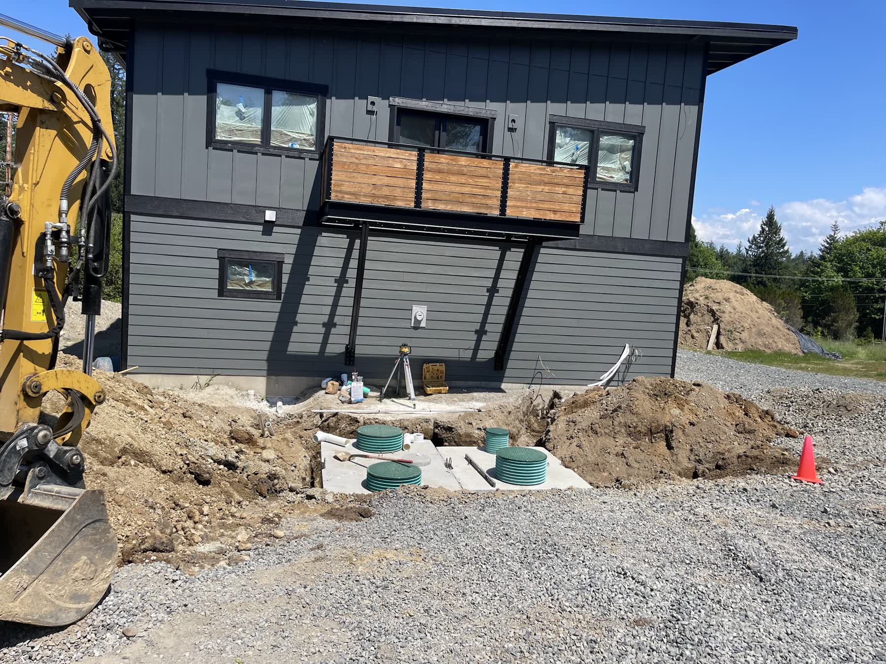 A new septic tank and risers being installed beside a newly built home, with a survey tripod and excavator on site.
