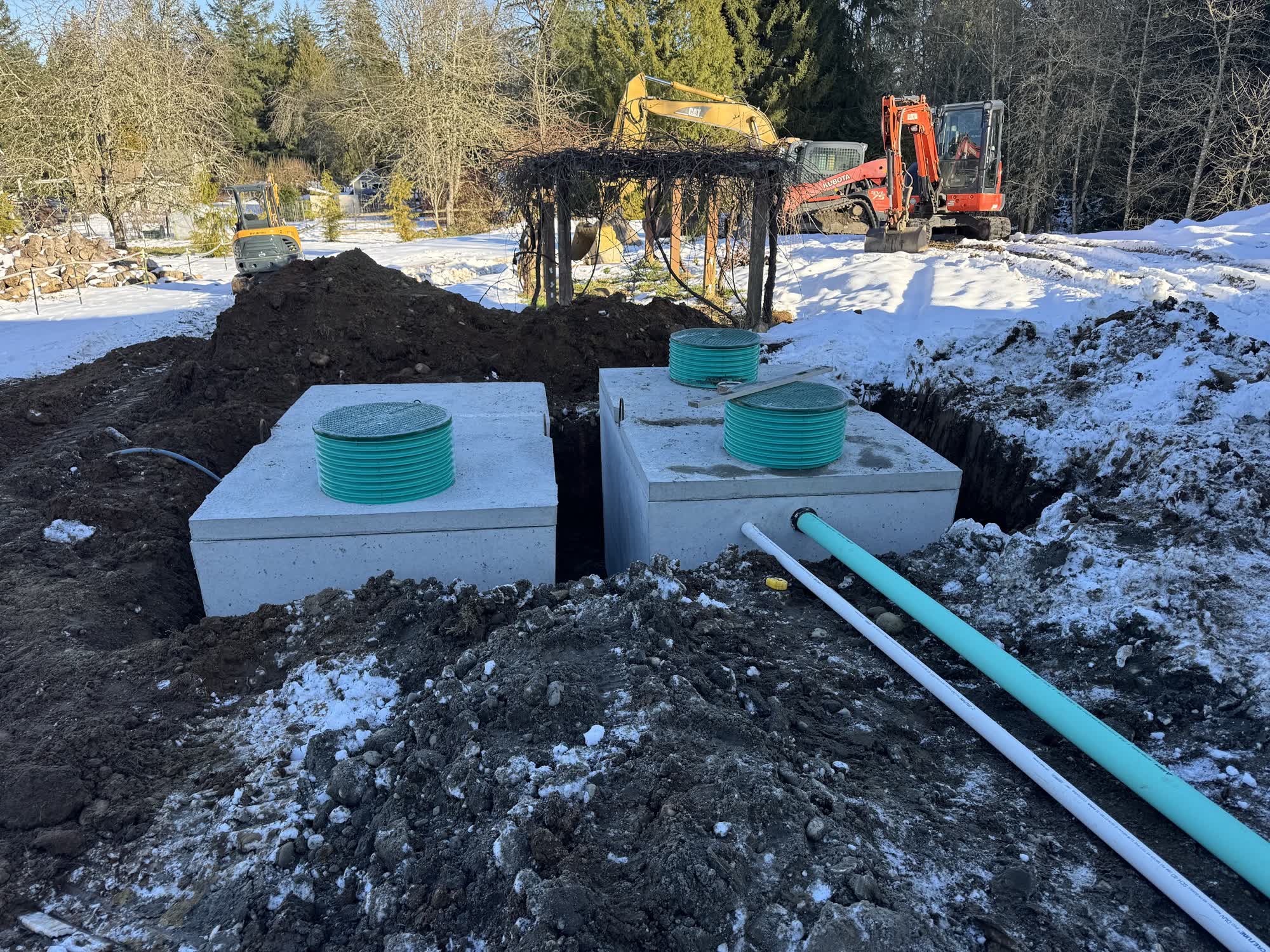 Two concrete septic tanks with teal risers set in the ground on a snowy site, with excavators working behind them.